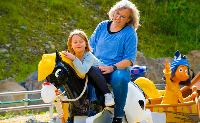 Oma und Enkelin genießen den Ausritt auf Kleiner Donner. Yakari winkt aus seinem Kanu.